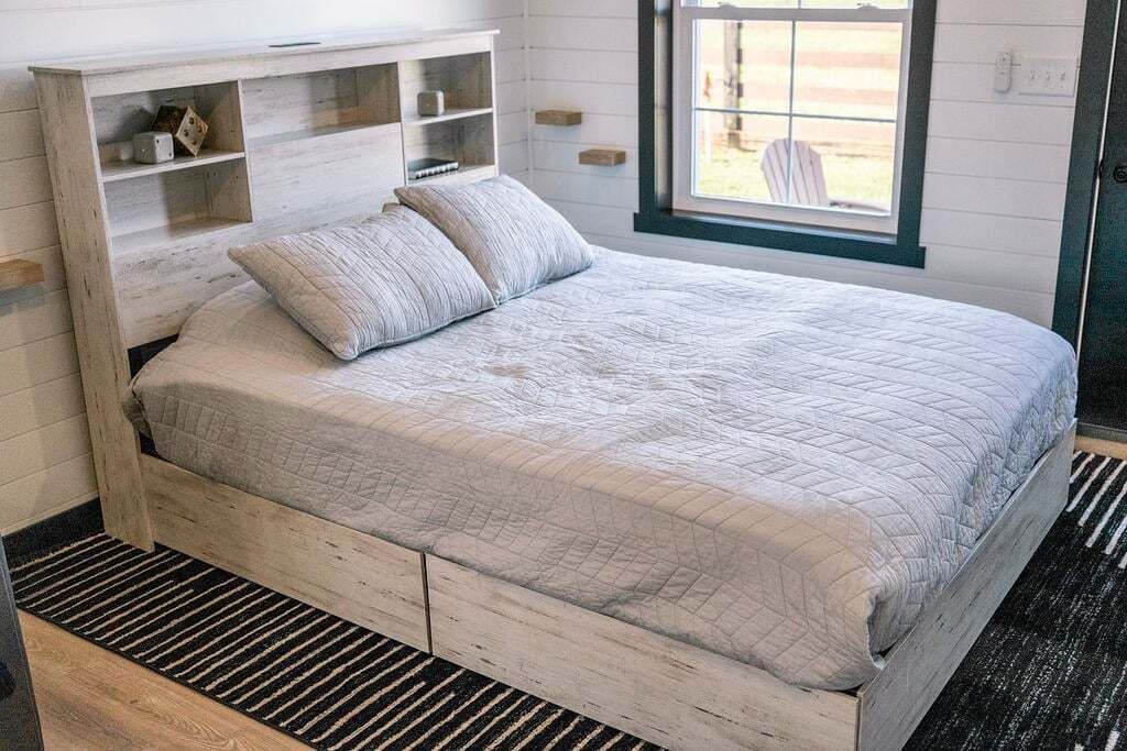 Ein Schlafzimmer mit einem Bett, einer Holzkommode mit offenen Regalen, einem Fenster mit Aussicht und einem Streifen Teppich.