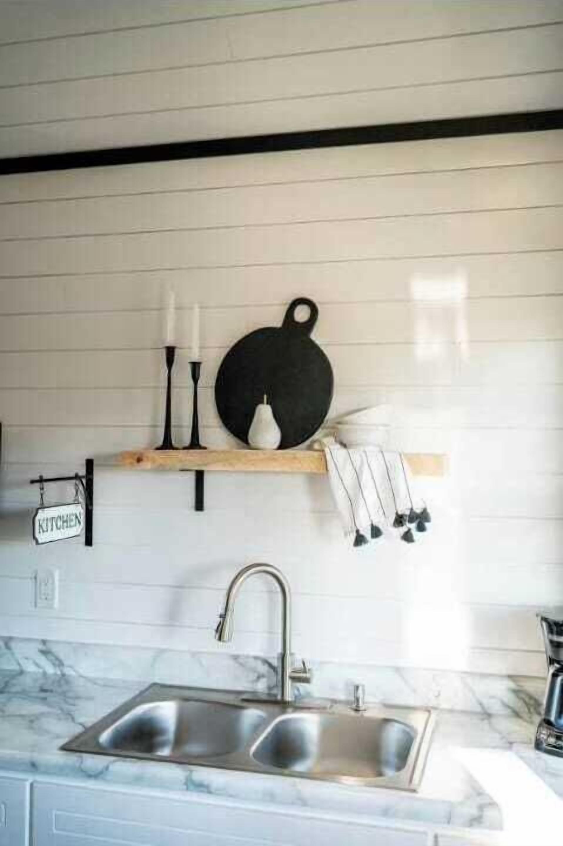 A kitchen with a marble countertop, a double sink, a hanging kitchen sign, and a shelf with candles and a pear.