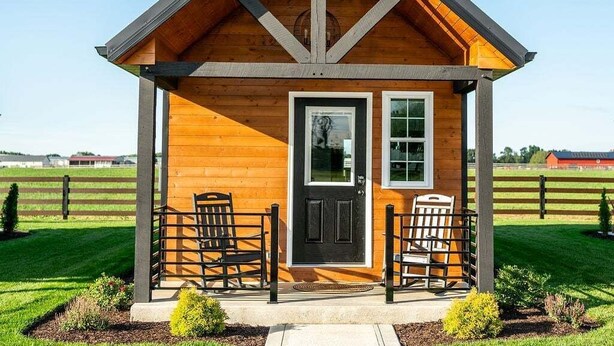 A small wooden cabin with a pitched roof, a front door, and two chairs outside.