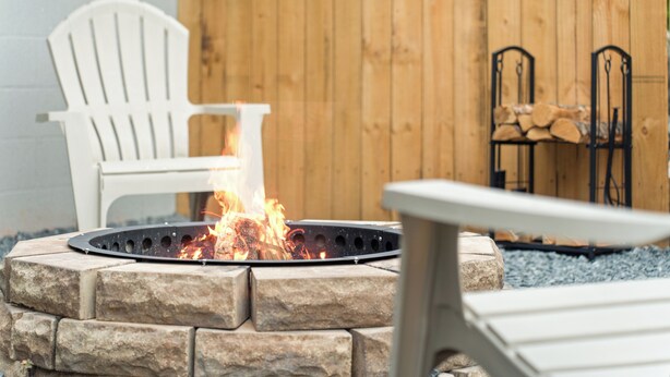 A stone fire pit with a metal grate, surrounded by a stone wall. There are two Adirondack chairs and a metal bench in the background. - Game Room Retreat • Fire Pit • 5 Min to Downtown (Mount Dora)