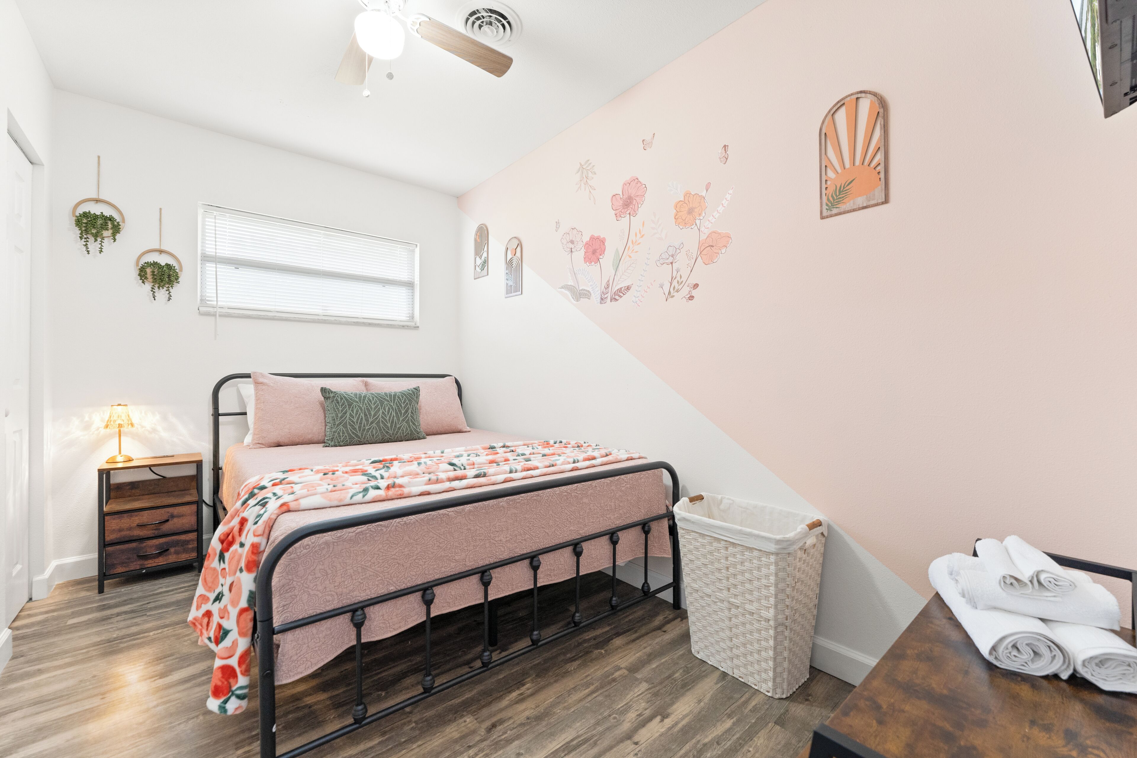 A bedroom with a bed, a nightstand, a lamp, a wall-mounted plant, a wall decoration, a trash bin, and a table with towels.