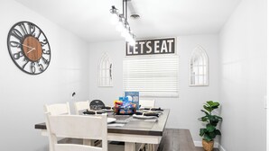 A dining area with a wooden table, white chairs, a clock, a 'LET'S EAT' sign, and a plant. - Game Room Retreat • Fire Pit • 5 Min to Downtown (Mount Dora)