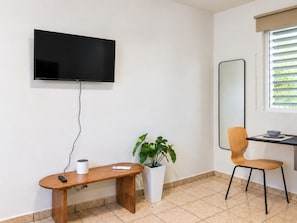 Ein modernes Zimmer mit einem wandmontierten Flachbildfernseher, einer Holzlounge mit Fernbedienung und einem weißen Behälter, einer Topfpflanze, einem Esstisch mit Stuhl und einem Fenster mit Jalousien.