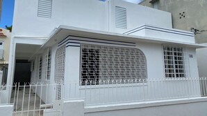A white two-story building with a covered front porch, a gated entrance, and a small window with a metal grid. - Cozy Studio 2@Casa Marvella / Water Tank (San Juan)