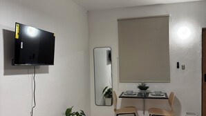 A room with a ceiling fan, a flat-screen TV mounted on the wall, a wooden bench, two chairs, a small table with a bowl and a plant, and a mirror on the wall. - Cozy Studio 2@Casa Marvella / Water Tank (San Juan)