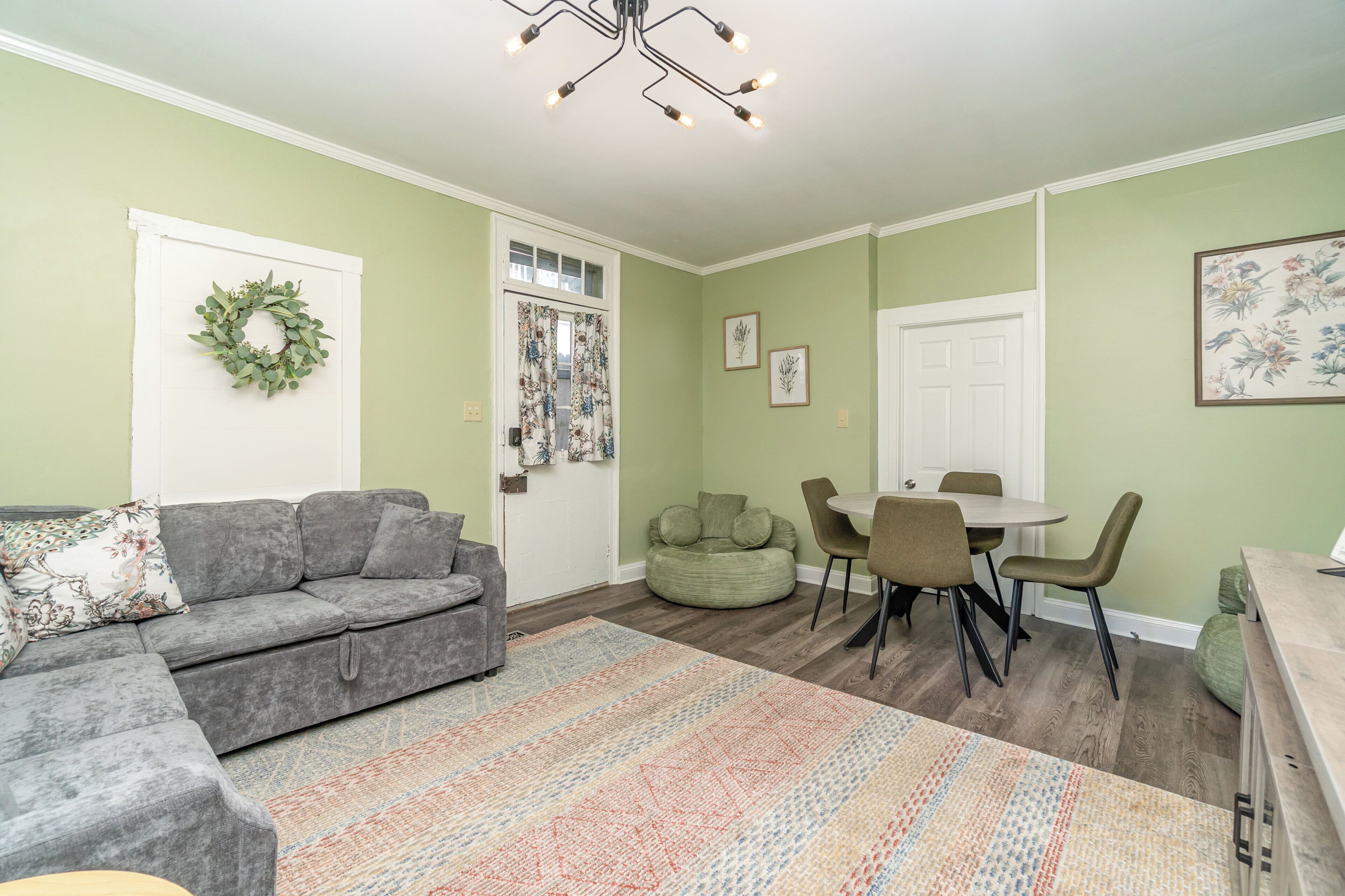 A living room with a grey sofa, a dining table with chairs, a green bean bag, and a colorful rug.