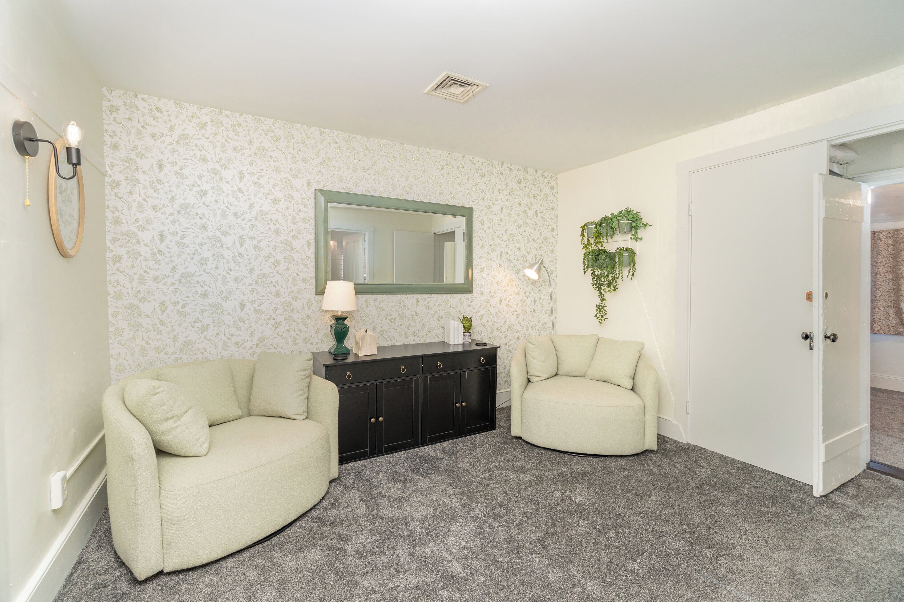A room with two cream-colored armchairs, a black cabinet with a mirror, and a door leading to another room.