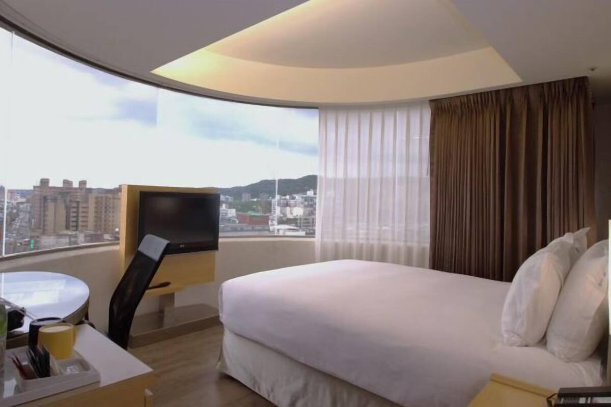 A hotel room with a bed, a TV, a desk with a chair, and a window with curtains.