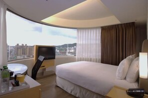 Habitación de hotel con cama, escritorio, silla, televisión y vista panorámica de la ciudad.
