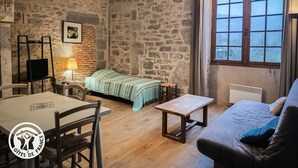 Una habitación rústica con pared de piedra, cama con manta verde, mesa de madera, sofá y ventana con vistas.