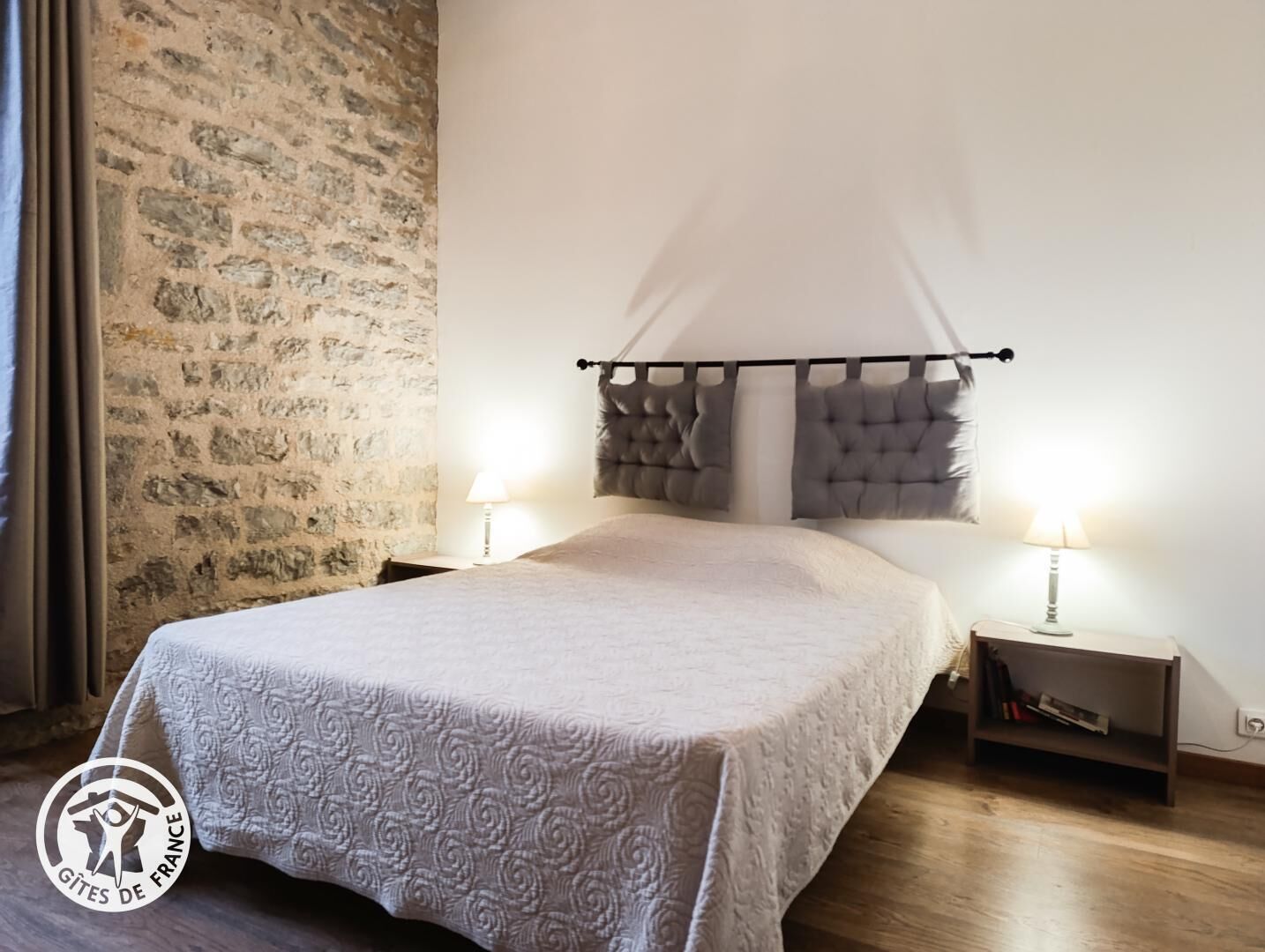 A bedroom with a stone wall, a bed with a tufted headboard, two bedside tables with lamps, and a wooden floor.