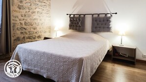 Un dormitorio con pared de piedra, una cama con cabecera capitonada, dos mesitas de noche con lámparas y suelo de madera.