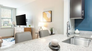 A modern hotel room with a kitchen area, a flat-screen TV mounted on the wall, a dining table with chairs, and a sink with a modern faucet. - Landing Furnished Apartments South End Stratford (Stratford)