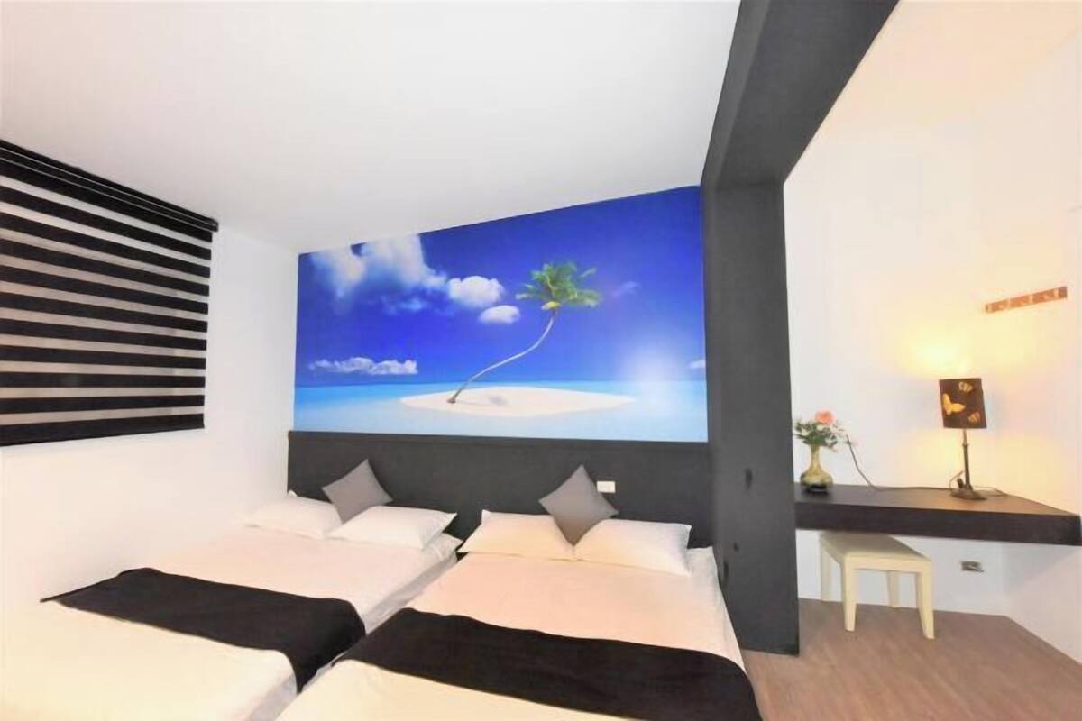 A bedroom with two beds, a large wall mural of a beach scene, a bedside table with a lamp, and a small stool.