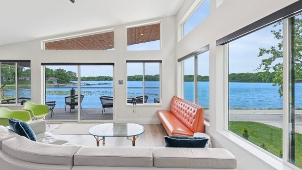 A spacious living room with a large window overlooking a lake, a white sofa, a glass coffee table, and a red bench. - New! Modern lakeside retreat-hot tub-trails-kayak (La Porte)