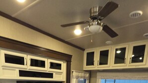 Cocina amplia en una caravana con un televisor grande, armarios blancos, ventilador de techo y sofá de cuero.