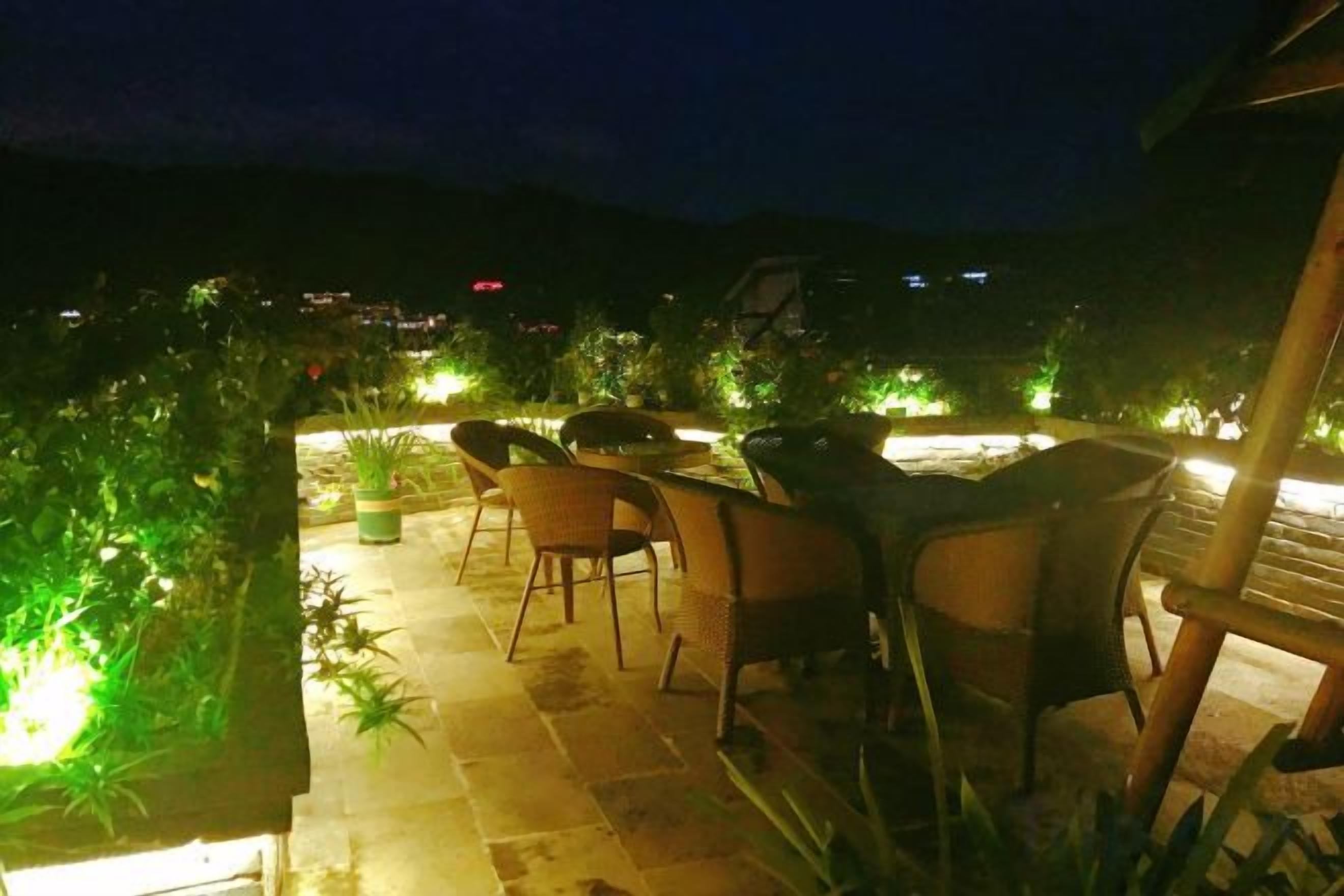 Un patio exterior con sillas y una mesa, rodeado de plantas en macetas e iluminado con luz ambiental.