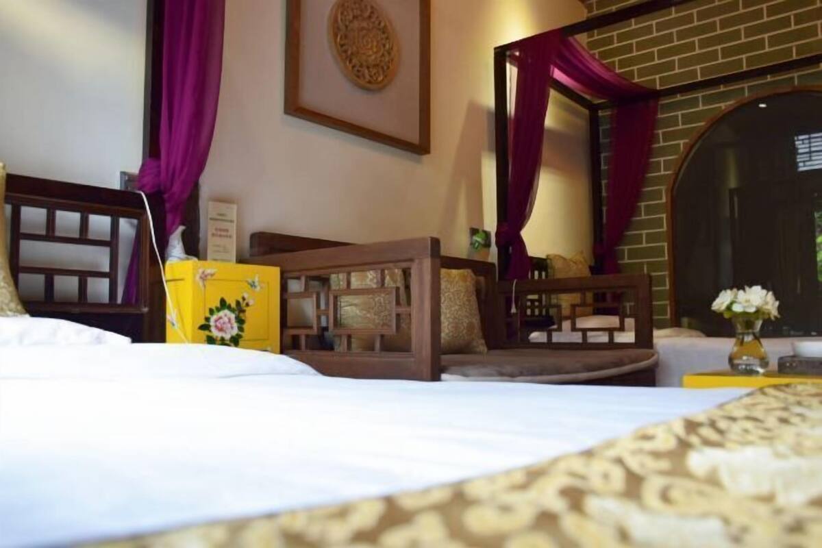 A hotel room with a bed, a wooden headboard, a small table with a vase of flowers, and a brick wall with a decorative mirror.