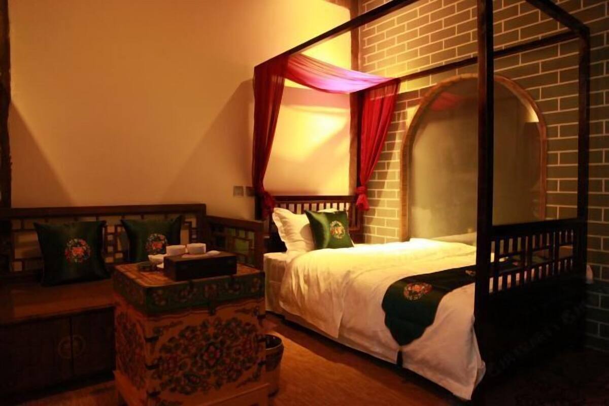 A room with a four-poster bed, a wooden chest of drawers, and a small table with a lamp.
