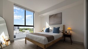 A modern bedroom with a large window, a bed with white bedding and blue pillows, a bedside lamp, and a view of the city. - Sleep 6 in Resort Style Community- Near Stadium (North Miami)