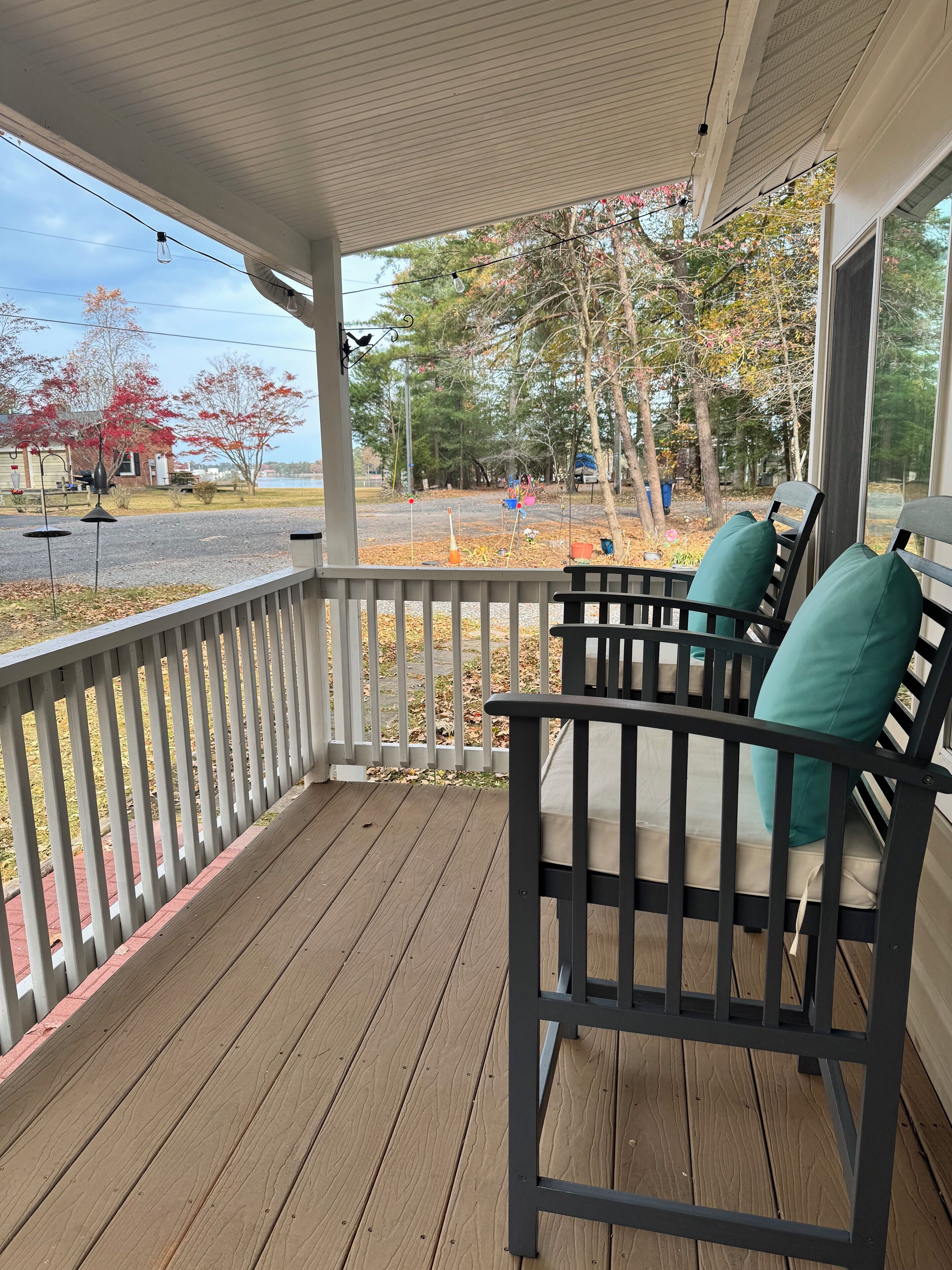 A wooden porch with a railing, two black chairs with teal cushions, and a view of a road and trees.