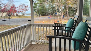 Une véranda en bois avec une balustrade, deux chaises noires avec des coussins turquoise, et une vue sur une route et des arbres.
