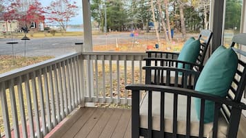 A wooden porch with a railing, two black chairs with teal cushions, and a view of a road and trees.