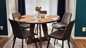 Un coin repas comprenant une table ronde, quatre chaises, un luminaire suspendu et une vue dégagée sur l’extérieur grâce à de grandes fenêtres ornées de rideaux.