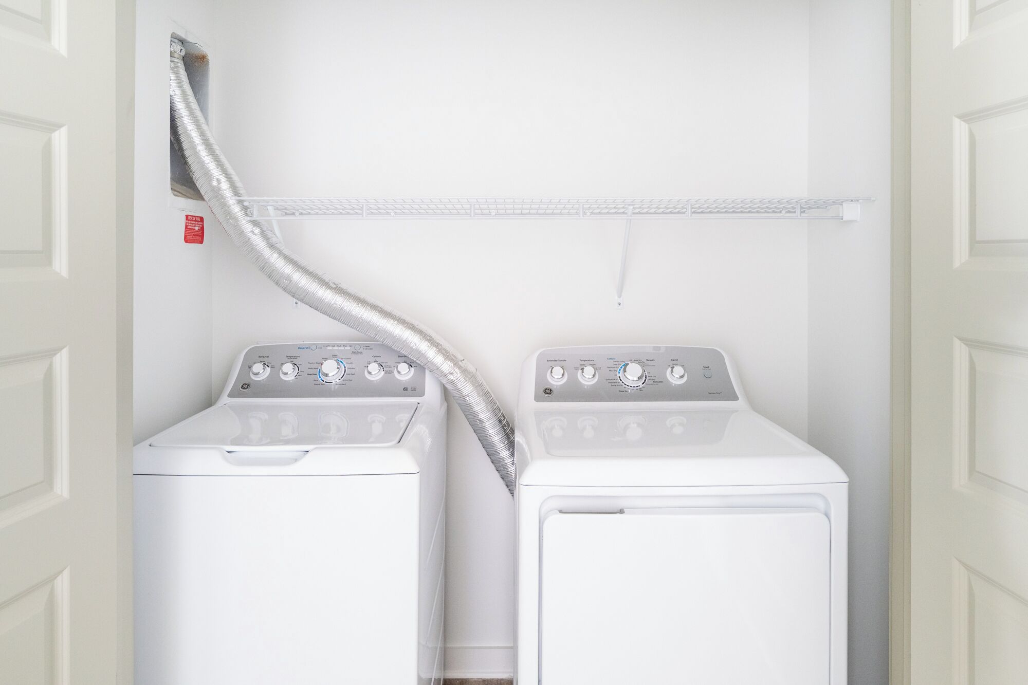A laundry room with two stacked front-loading washers and dryers, a metal vent, and a white shelving unit above.