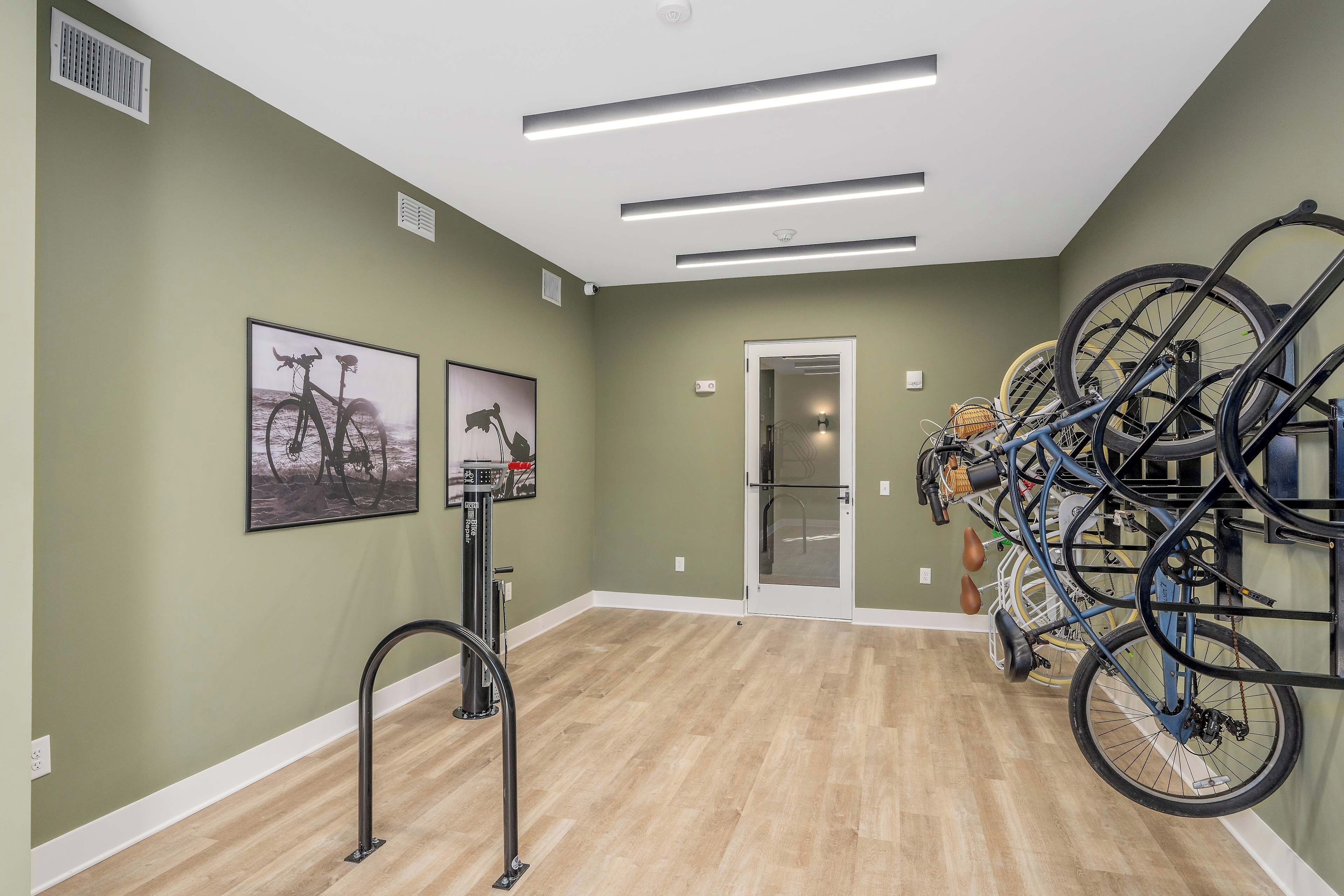 A room with green walls, wooden flooring, and a door leading to another area. There are bicycles mounted on the wall and a bike rack with multiple bicycles.