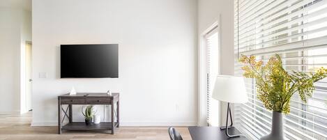 A modern living room with a flat-screen TV, a dark wooden coffee table, a black chair, a vase with flowers, and a window with blinds.