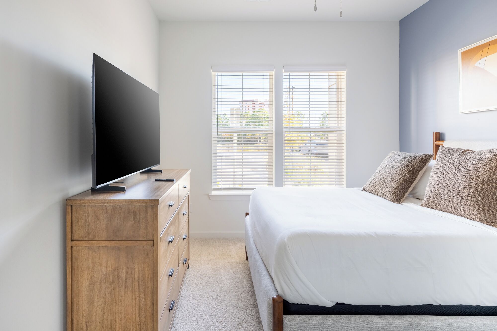 A bedroom with a bed, a wooden dresser, a flat-screen TV, and a window with blinds.
