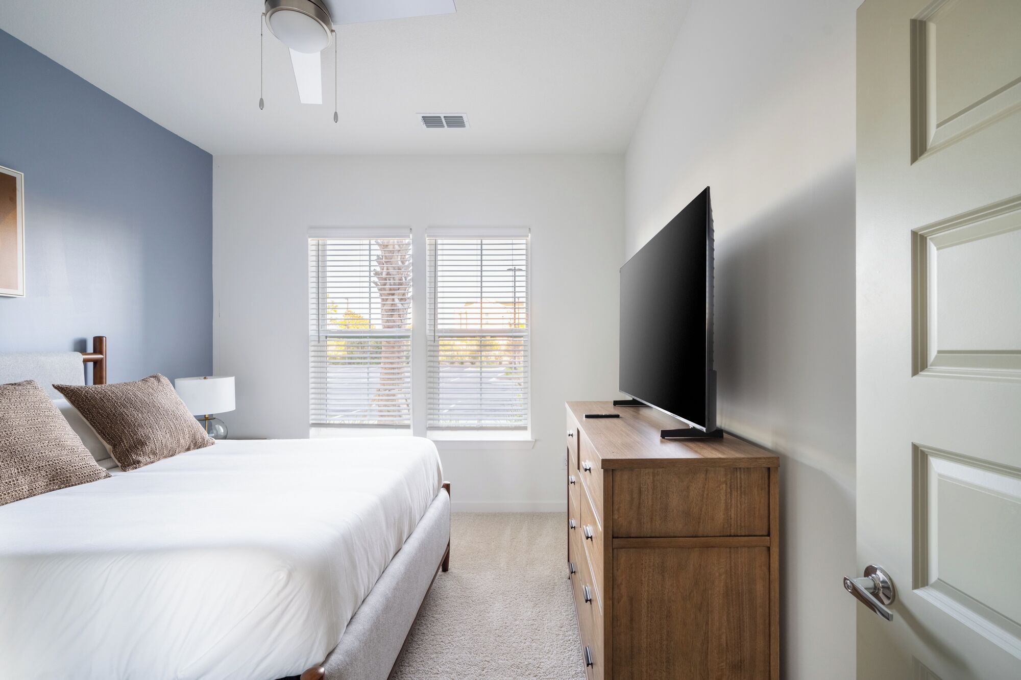 A bedroom with a bed, a TV on a wooden dresser, and a window with blinds.