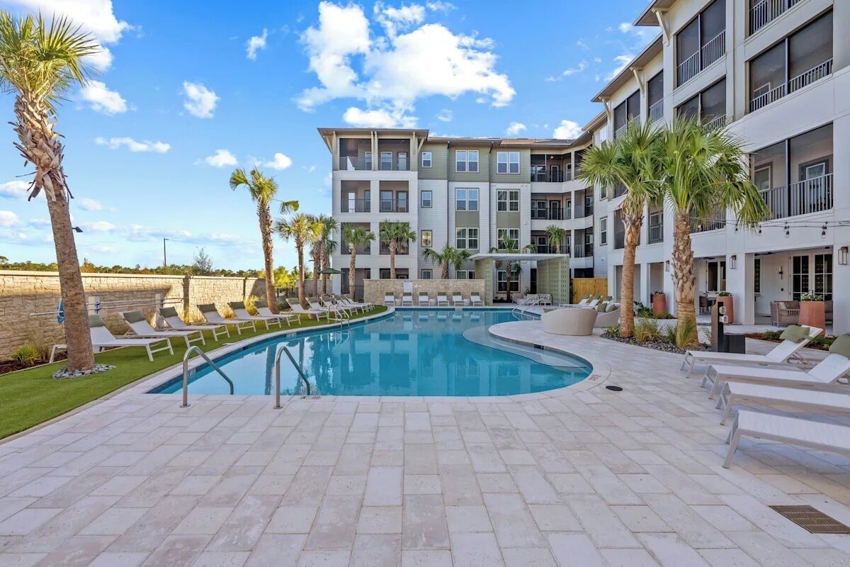 A pool area with lounge chairs and palm trees, surrounded by a multi-story building with balconies.