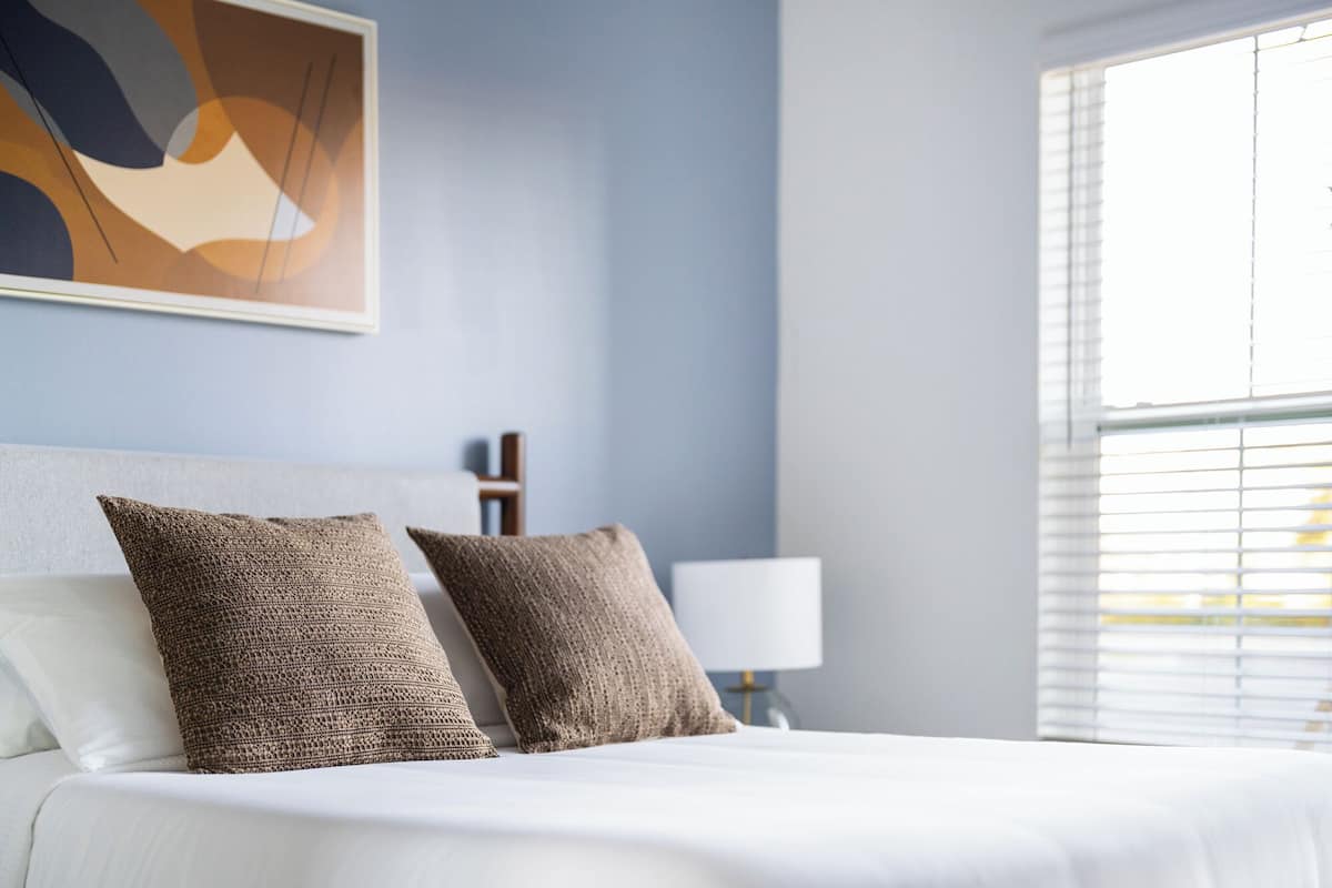 A neatly made bed with two textured pillows, a bedside table with a lamp, and a window with blinds.