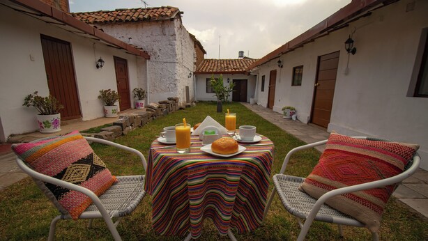 An outdoor dining area with a table set for two, surrounded by chairs, in a courtyard with a white building in the background. - Encanto San Blas (Cusco)