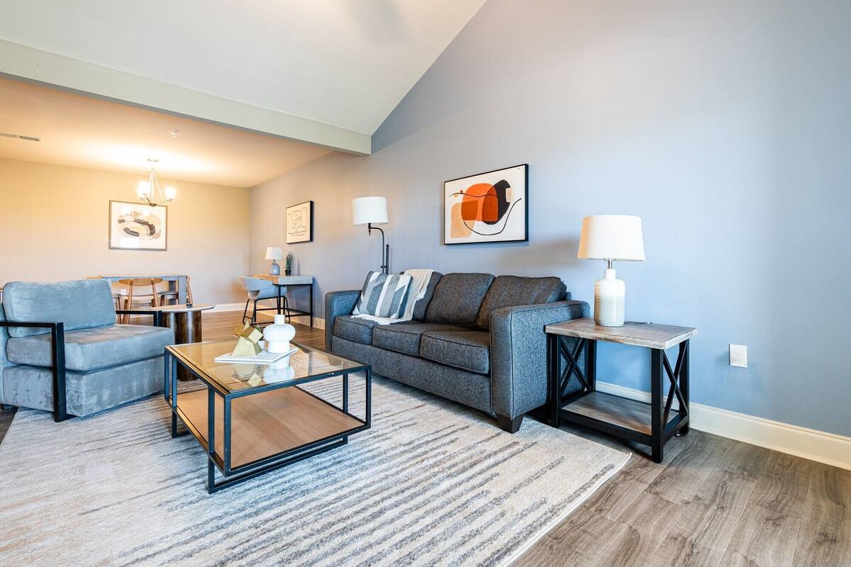 Een moderne woonkamer met een grijze bank, een blauwe fauteuil, een glazen salontafel en een bijzettafeltje met een lamp.