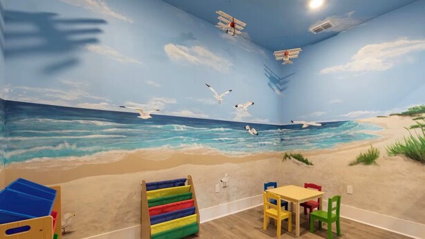 Une pièce décorée d’une fresque représentant une plage, une cabane en bois servant d’aire de jeux, une table avec des chaises et un meuble de rangement coloré.