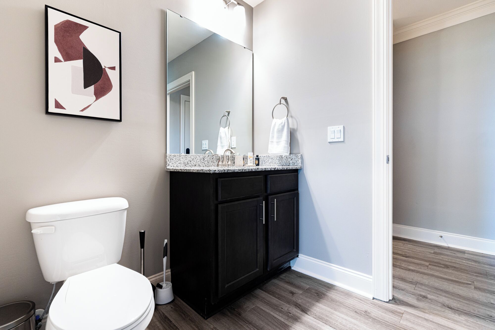 A bathroom with a toilet, dark cabinets, a granite countertop, and a mirror.