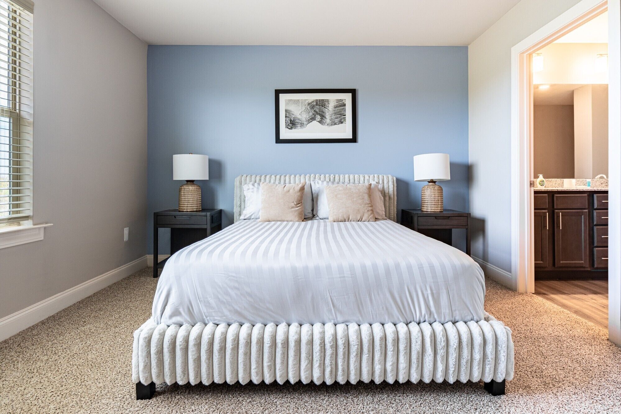 A bedroom with a large bed, two bedside tables with lamps, a framed artwork on the wall, and a view into the bathroom.
