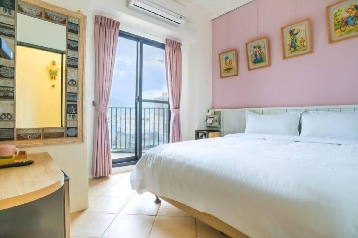 Un dormitorio con una cama grande, un escritorio con silla, una ventana con cortinas y un balcón.
