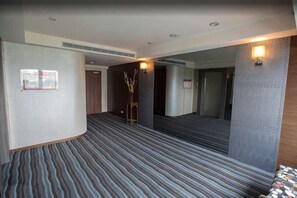Quarto de hotel com carpete, uma cama, uma mesinha de cabeceira, uma luminária na parede e uma porta.