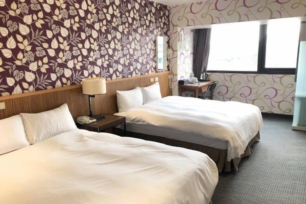 A hotel room with two beds, a nightstand, a lamp, a desk, and a window with curtains.