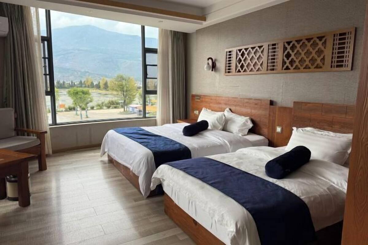 Une chambre d’hôtel avec deux lits, une tête de lit en bois, une fenêtre donnant sur la montagne et une petite table avec une chaise.