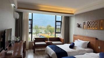 Habitación de hotel con dos camas, televisión, escritorio y ventana grande con vista a árboles y montañas.