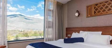 A hotel room with a large window offering a mountain view, a wooden headboard, and a decorative wall panel.