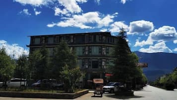A multi-story building with a parking area in front, surrounded by trees and mountains in the background.