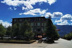 A multi-story building with a parking area in front, surrounded by trees and mountains in the background. - CHP. Hotel (Heqing)