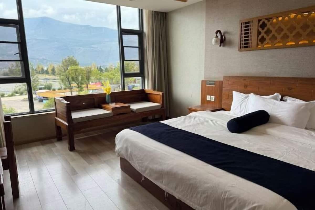 Une chambre d’hôtel avec un lit, un banc en bois, une table de chevet et une vue sur les montagnes par de grandes fenêtres.