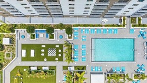 An aerial view of a modern hotel with multiple swimming pools, sun loungers, and a well-maintained garden area.